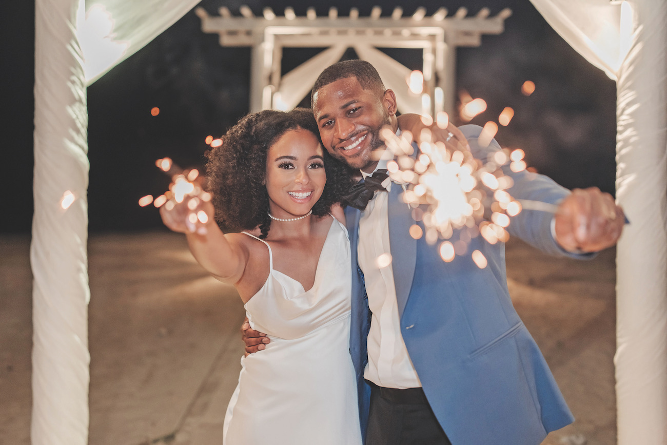 A couple dressed formally holds sparklers and smiles at the camera under a decorated outdoor structure at night.