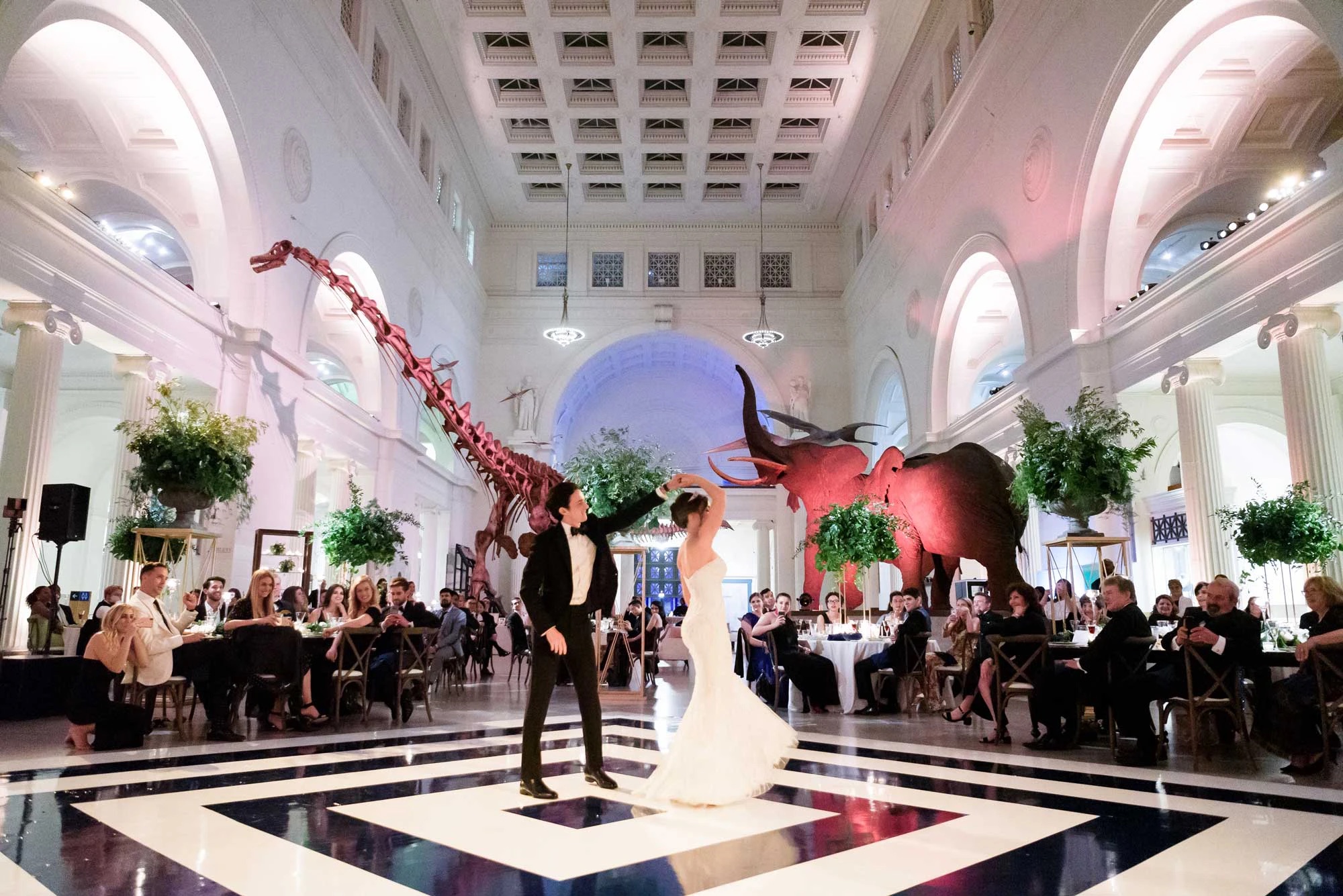 A bride and groom share their first dance in a grand hall with dinosaur and mammoth exhibits, surrounded by seated guests.