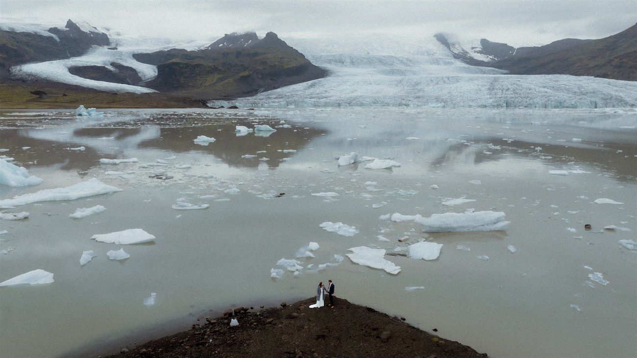 Two people stand on a rocky outcrop by a glacial lagoon with floating icebergs, mountains, and a glacier in the background under a cloudy sky.