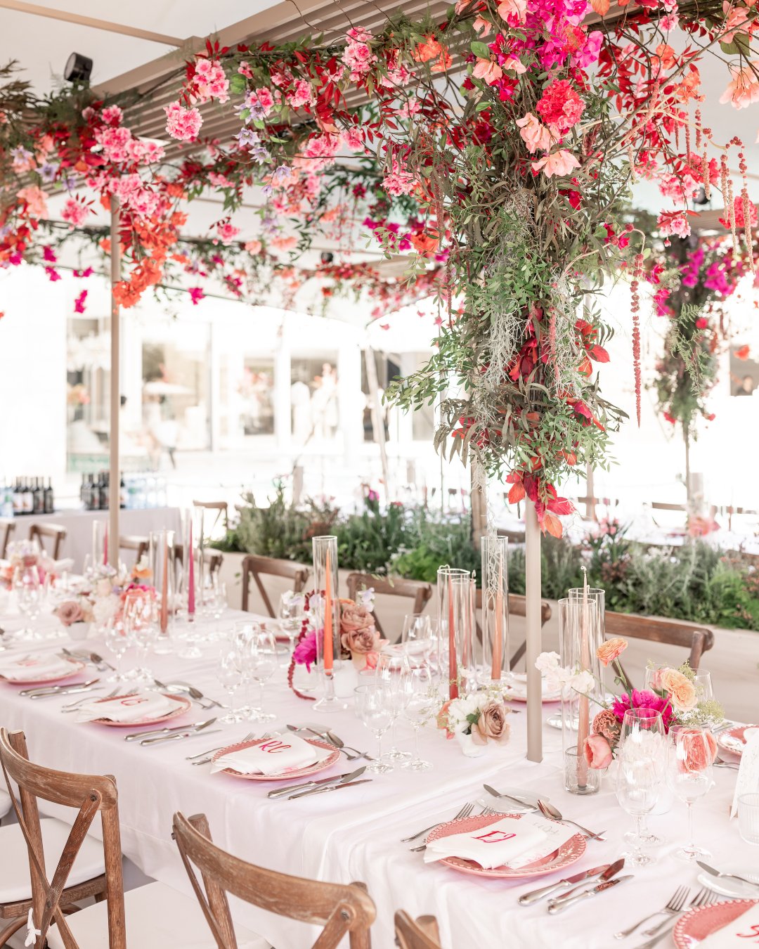 A long dining table with wooden chairs is set with white linens, pink-themed tableware, and decorated with hanging pink and red flowers.
