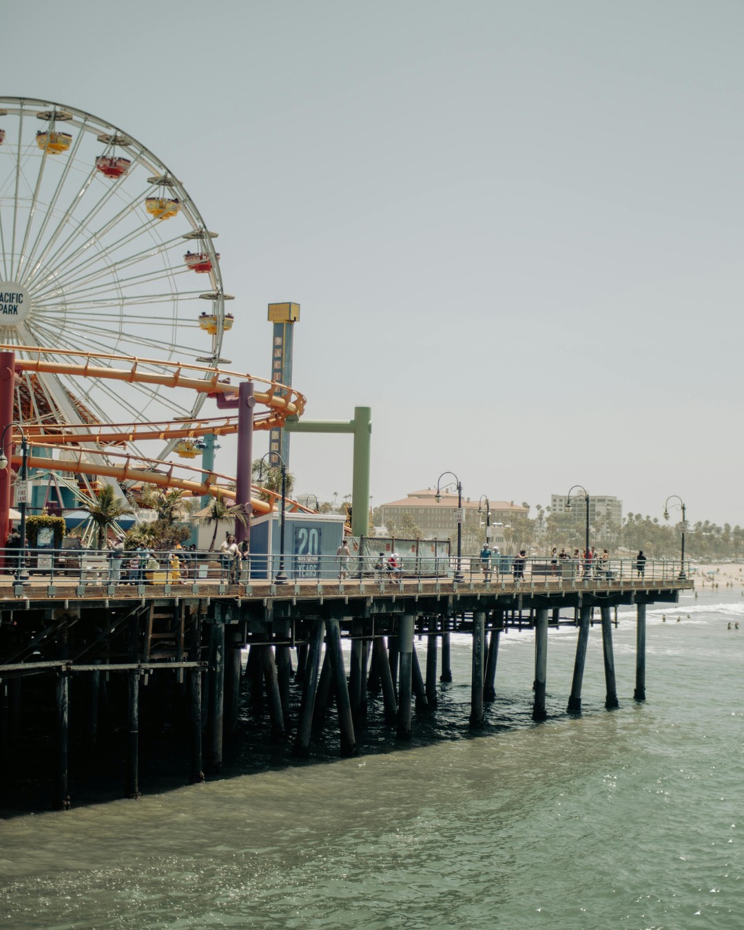 A large Ferris wheel and roller coaster on a wooden pier extend over the ocean, with people walking and the shore visible in the background.