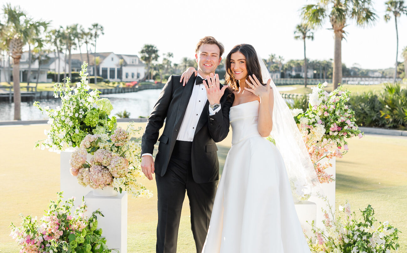 A bride and groom stand outdoors in front of floral arrangements, smiling and showing their wedding rings.