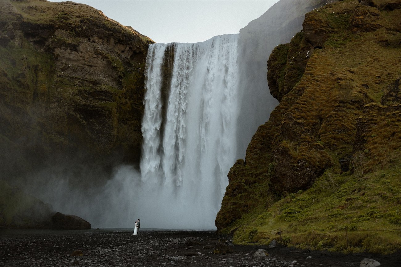 A person in a white outfit stands near the base of a large waterfall surrounded by mossy cliffs and mist.