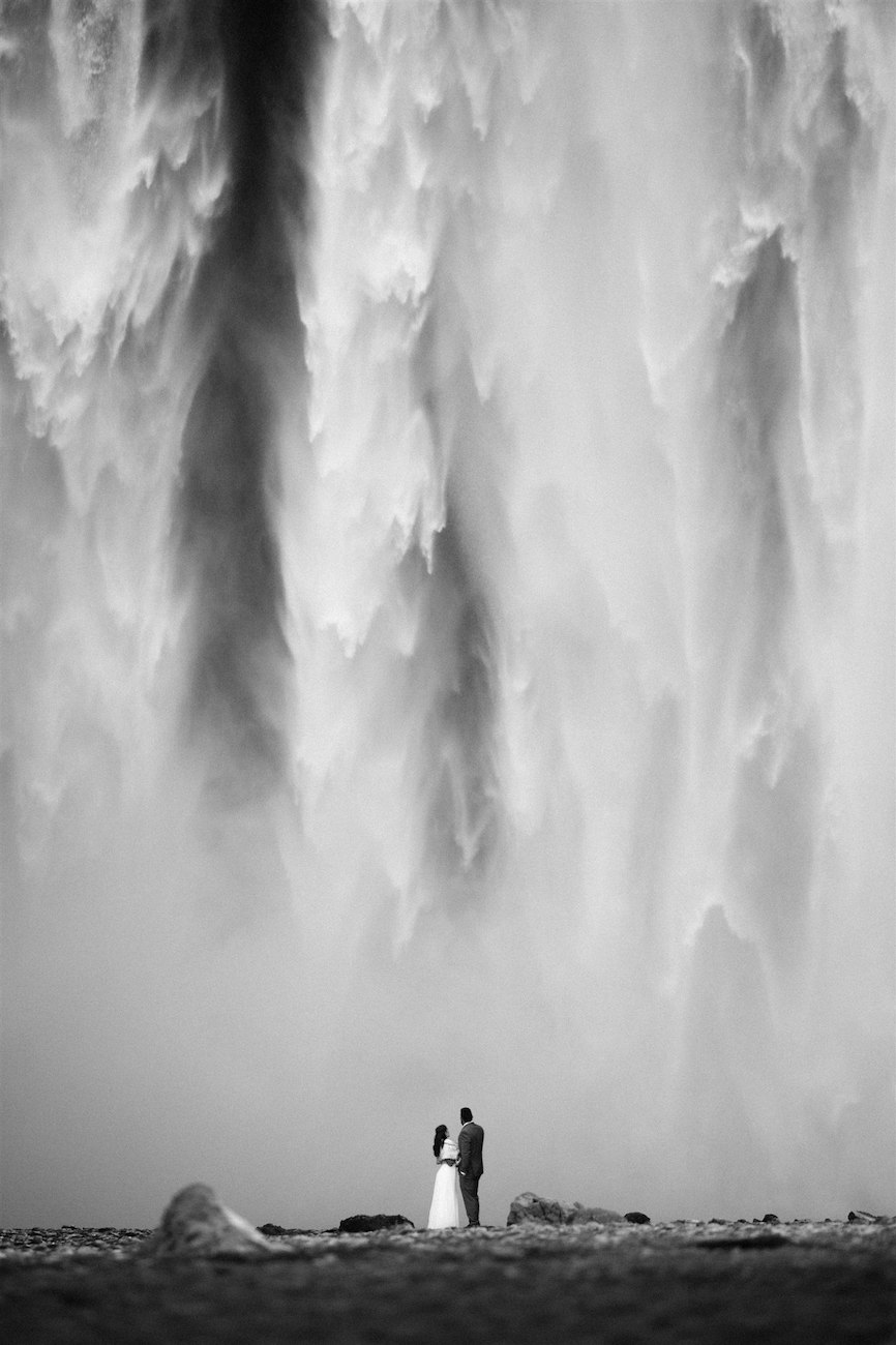 A couple stands together near the base of a large, dramatic waterfall, with mist and water cascading down behind them. Black and white photo.