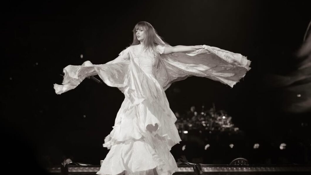 A woman in a flowing dress dances on stage with her arms extended, creating a dramatic, wing-like effect with the fabric. The image is in black and white.