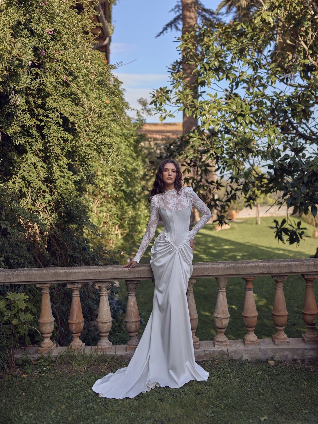 A woman in a long-sleeved, floor-length white gown stands by a stone balustrade in a lush garden with tall trees and greenery in the background.