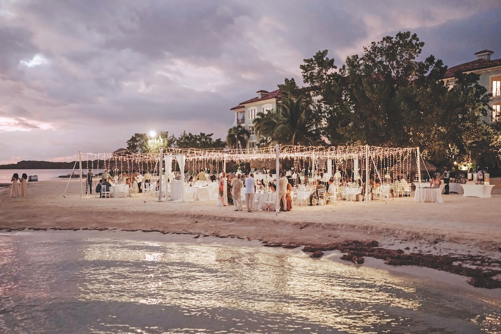 Sandals Resorts Wedding reception with tables and string lights set up on a sandy beach at dusk, with guests gathered and buildings and trees in the background.