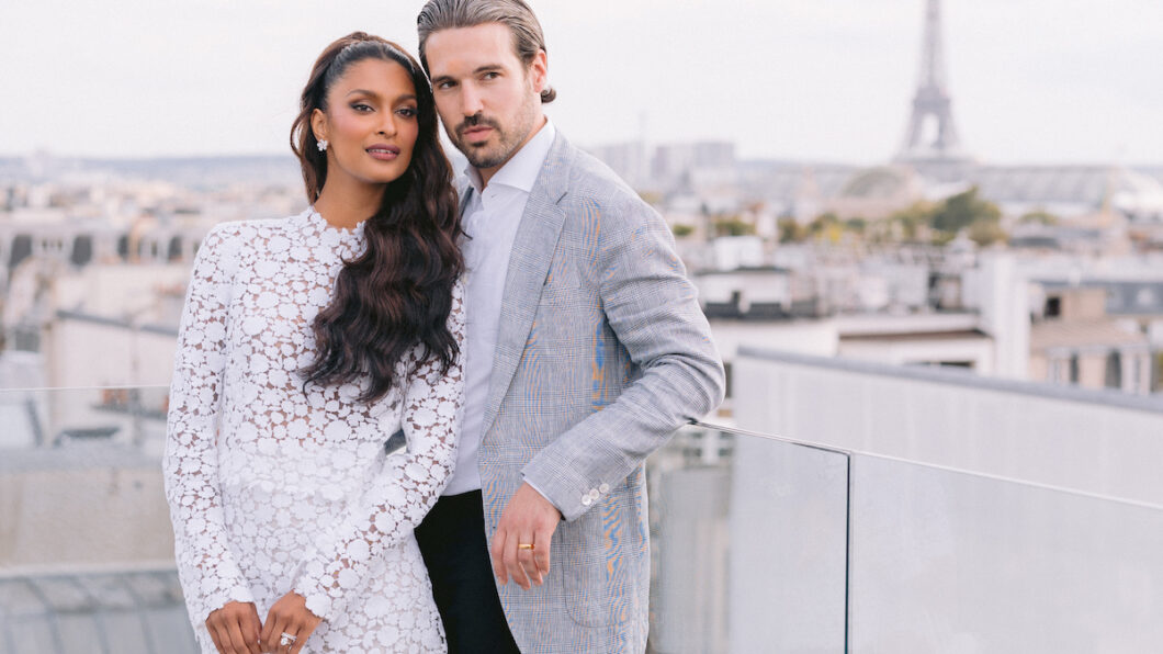 A woman in a white dress and a man in a grey blazer pose together on a rooftop with the Eiffel Tower visible in the background.