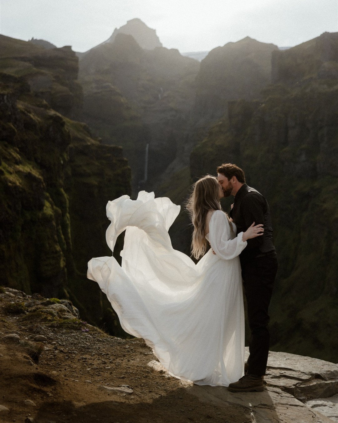 A couple stands on a rocky ledge, the woman in a flowing white dress and the man in dark clothing, embracing in front of a mountainous landscape.
