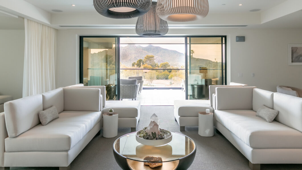 Modern living room with white sofas, round coffee table, and large pendant lights; glass doors reveal a patio with mountain views in the background.