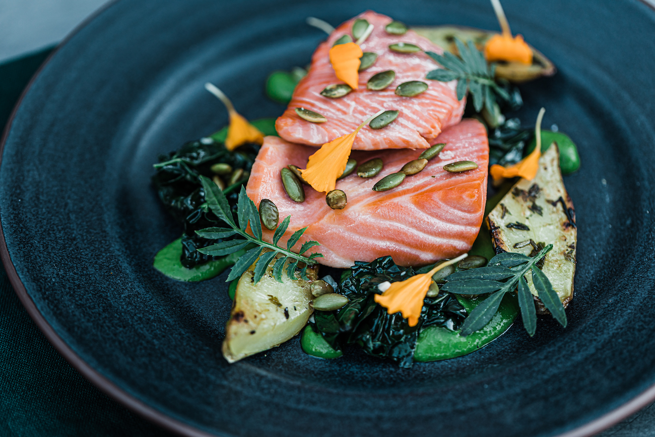 Two slices of raw salmon topped with pumpkin seeds, served on grilled vegetables, leafy greens, and garnished with yellow edible flowers on a dark plate.