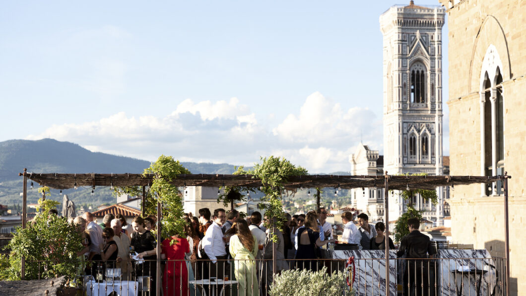 A group of people gather on a rooftop terrace with vines overhead, socializing with drinks. A tall stone tower stands in the background on a clear day.