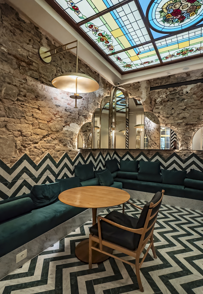A cozy lounge features zigzag patterned walls and floor, stone walls, green upholstered seating, a round wooden table, and a stained glass ceiling.