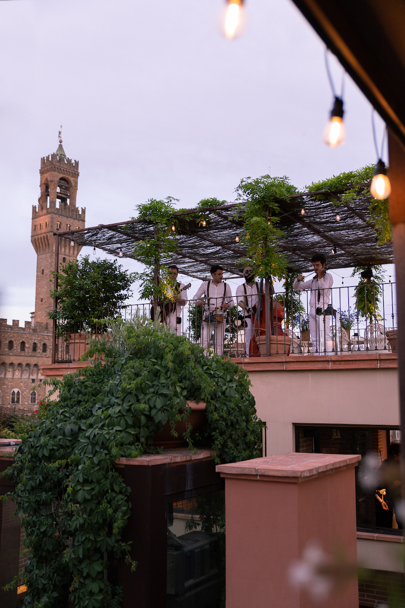 Musicians perform on a rooftop terrace covered with greenery, with a historic stone tower visible in the background under a cloudy sky.