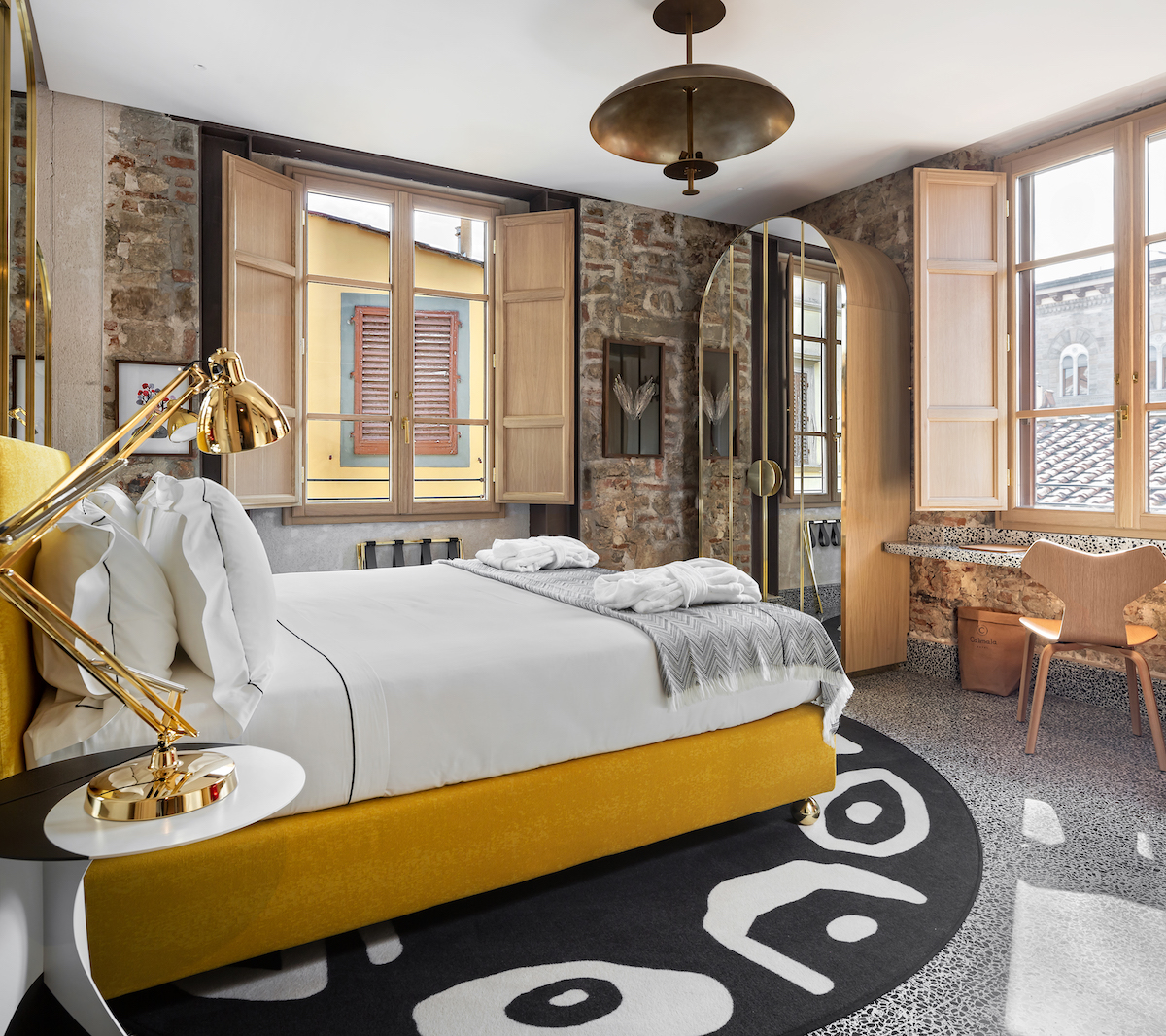 A stylish bedroom with exposed brick walls, large windows with wooden shutters, a yellow bed, a modern desk, and a black and white patterned rug.