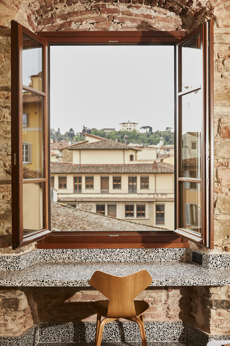 Open window in a brick wall above a granite desk with a wooden chair, overlooking rooftops and distant buildings.