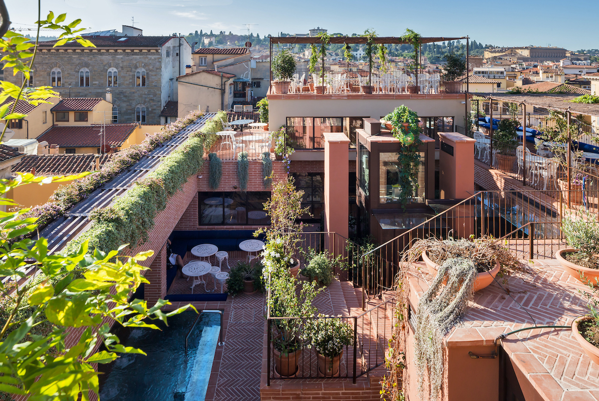 Multi-level rooftop terrace with plants, seating areas, and a small pool, overlooking a cityscape under a clear sky.