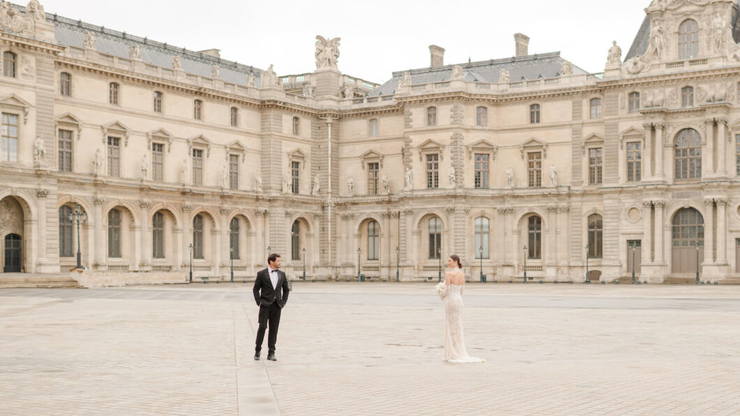 A man in a tuxedo and a woman in a white dress stand apart in an empty courtyard in front of a historic building. Featured image for Your Guide to Iconic Paris Wedding Photos