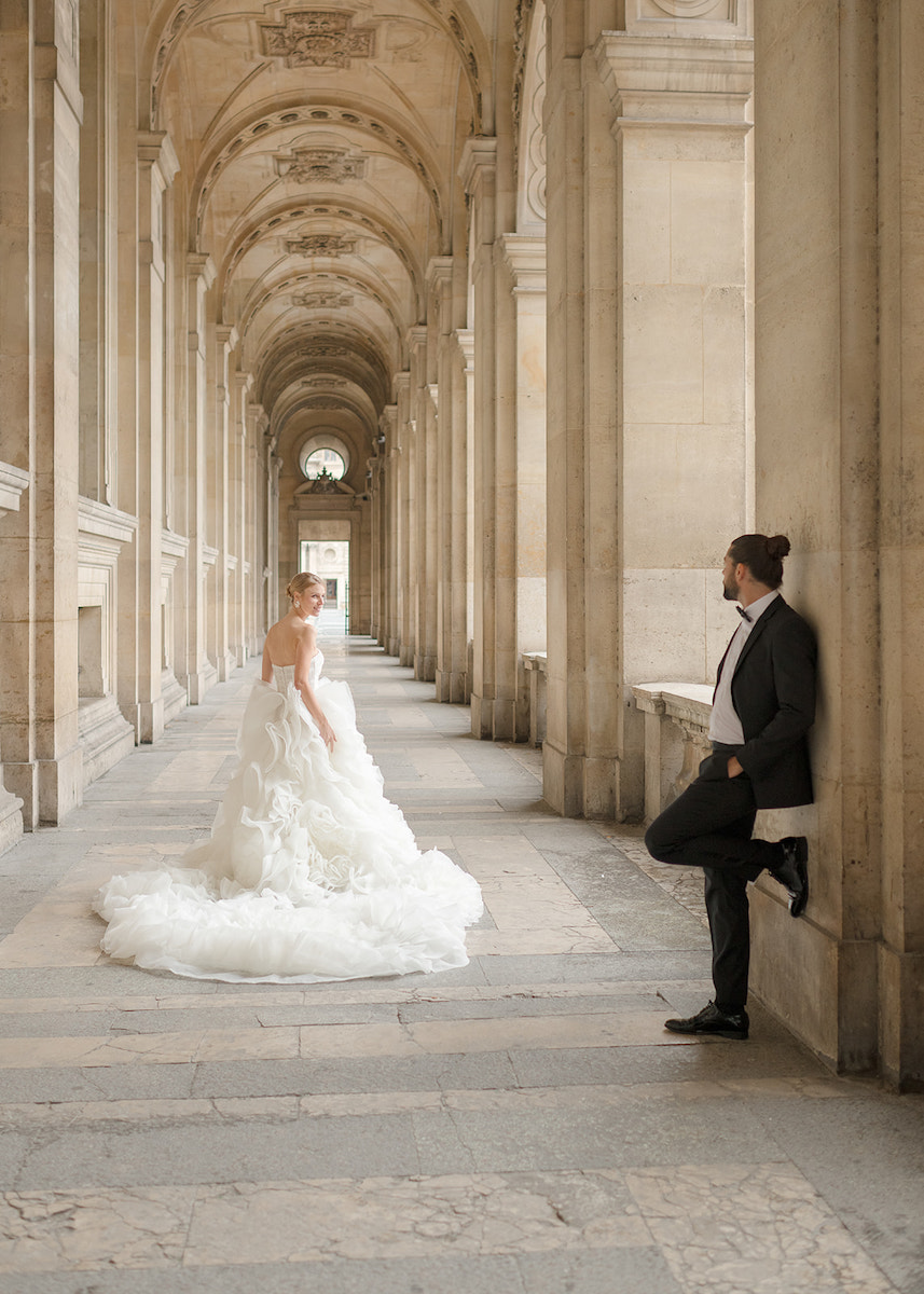 A bride in a white gown walks toward a man in a suit, who is leaning against a column in an ornate, arched hallway.
