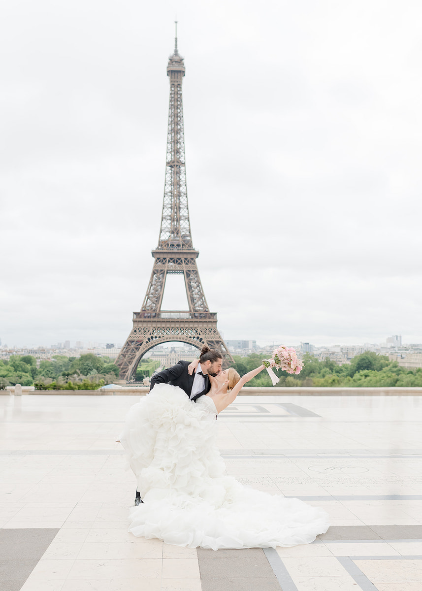 A bride and groom pose in a dip in front of the Eiffel Tower on a cloudy day, with the bride holding a bouquet aloft.