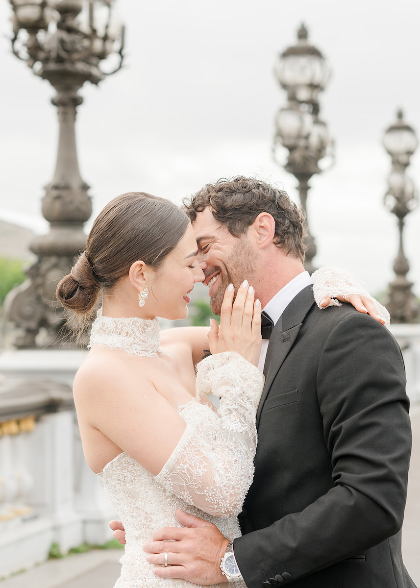 A bride and groom embrace and smile at each other on an ornate bridge with decorative street lamps in the background.