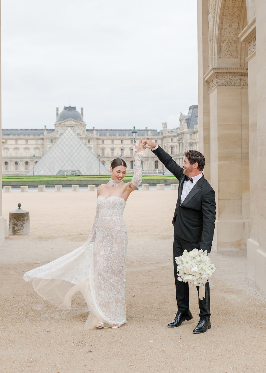 A bride and groom pose together outside the Louvre Museum, with the groom twirling the bride while holding a bouquet of white flowers.