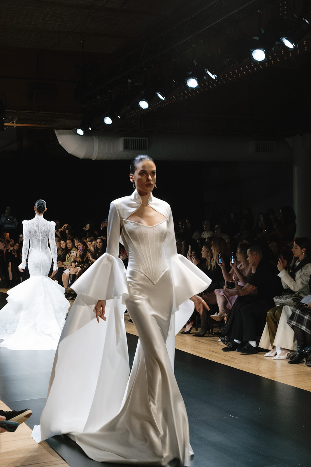 A model walks down a runway wearing a long, white, fitted gown with dramatic sleeves; audience members are seated on both sides.
