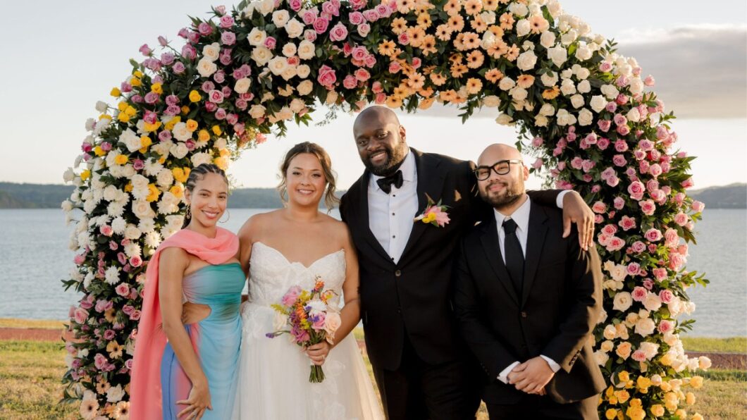 Four people stand smiling under a large floral arch; one in a wedding dress, one in a tuxedo, and two in formal attire.