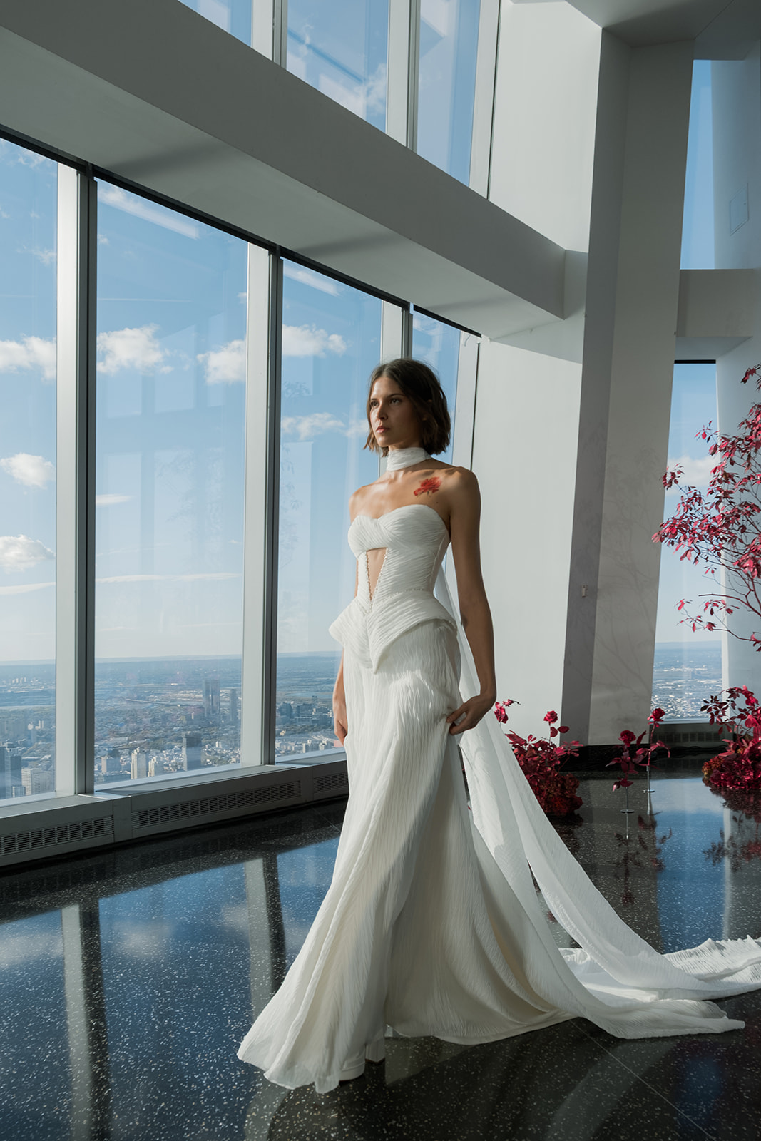 A woman in a modern white wedding dress stands by large floor-to-ceiling windows with city views and pink floral arrangements nearby.