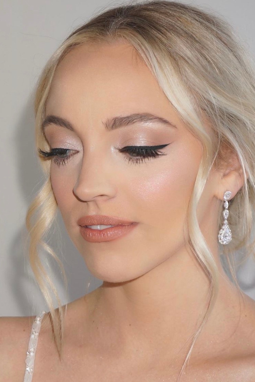 A woman with blonde hair and light skin wears dramatic winged eyeliner, nude lipstick, and drop earrings, looking down with her eyes closed.