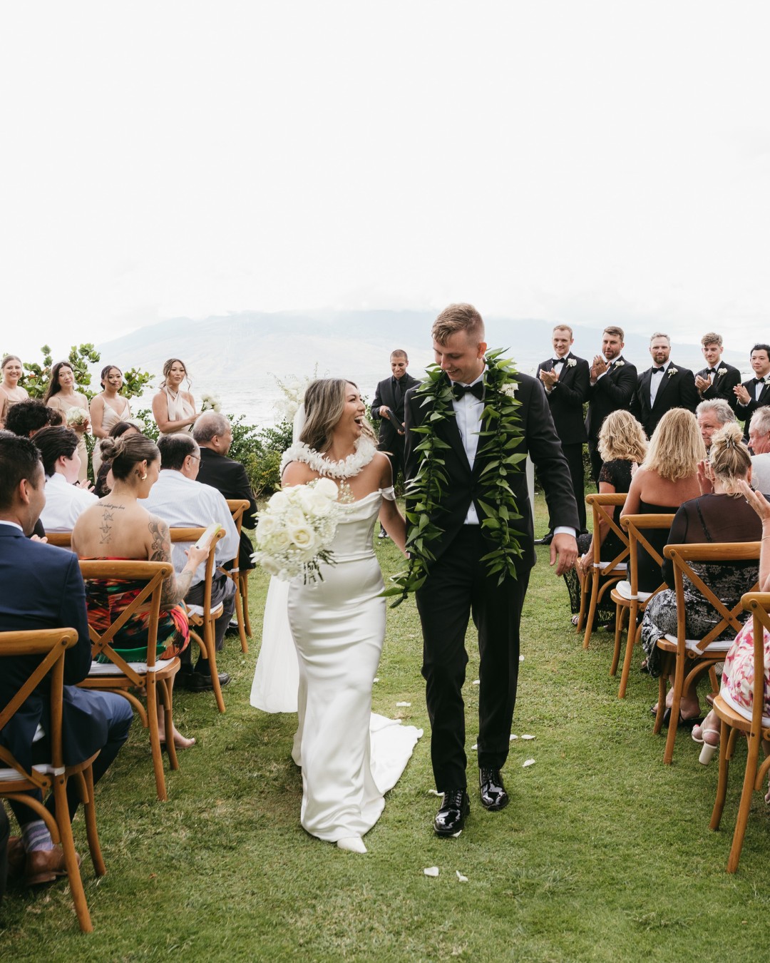 A bride and groom walk down the aisle outdoors, surrounded by seated guests and a wedding party dressed in black, with greenery and mountains in the background.