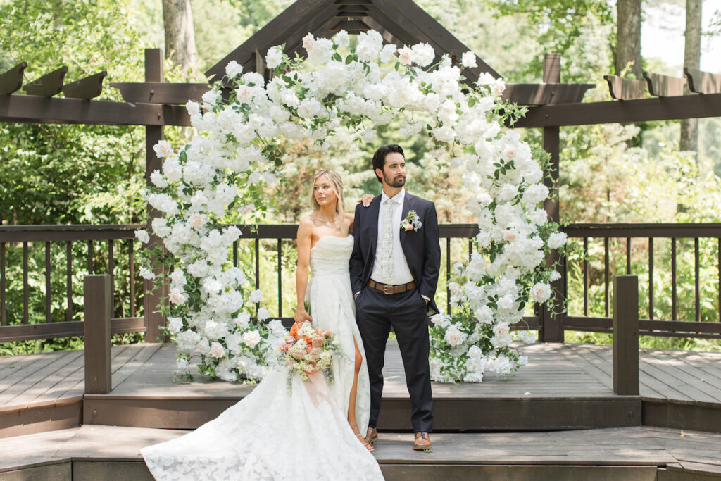 A bride and groom stand side by side on a wooden platform, in front of a large circular floral arrangement outdoors.