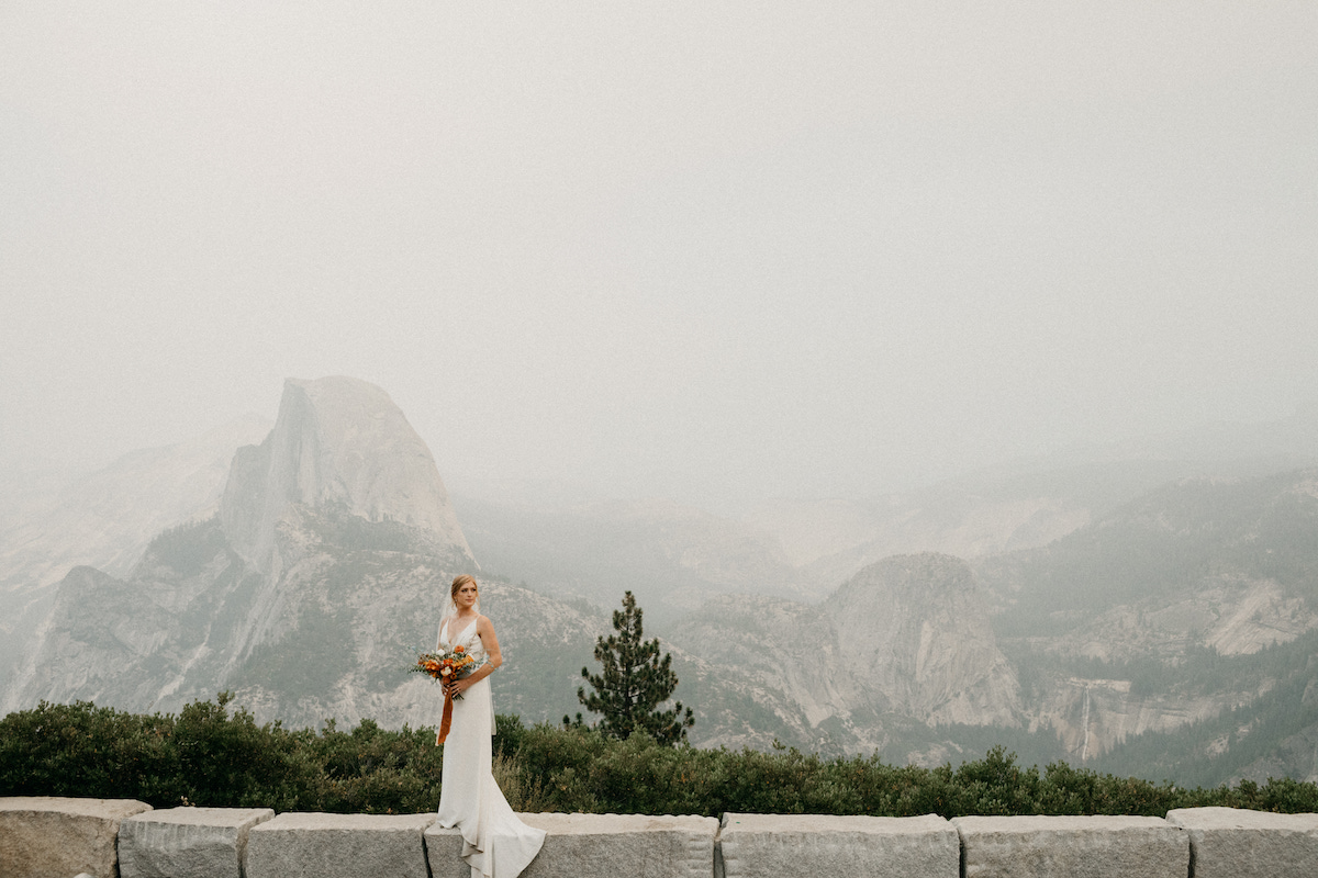 A bride in a white dress holding a bouquet stands on a stone ledge with a mountainous landscape and Half Dome in the background.