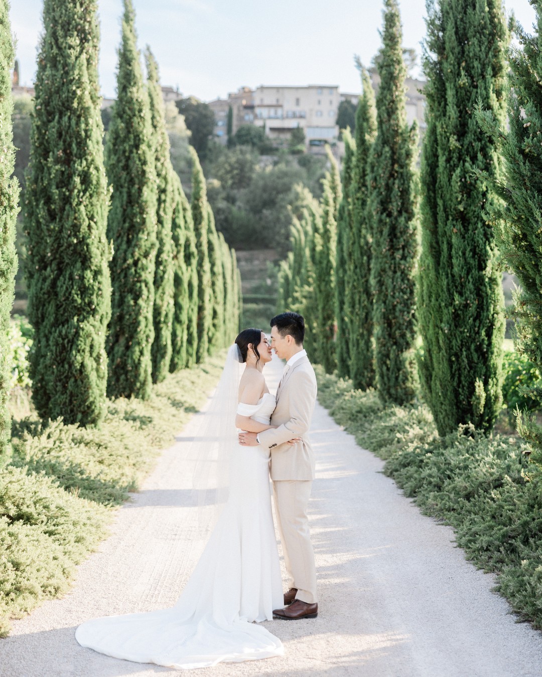 A bride and groom stand embracing on a tree-lined path with tall cypress trees and distant buildings in the background.