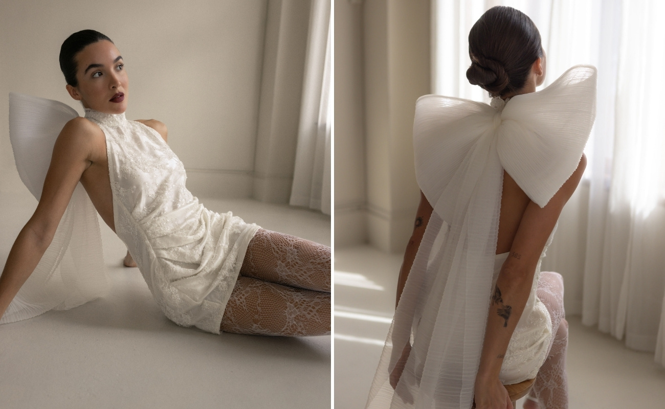 A woman in a sleeveless, short white dress featuring a large sheer bow on the back sits and kneels on the floor in natural light.