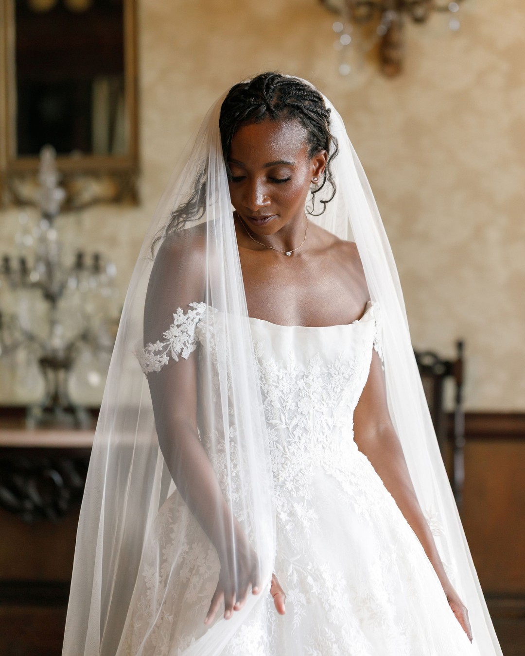 A bride in a white, off-the-shoulder wedding dress with lace details and a long veil stands indoors, looking down.