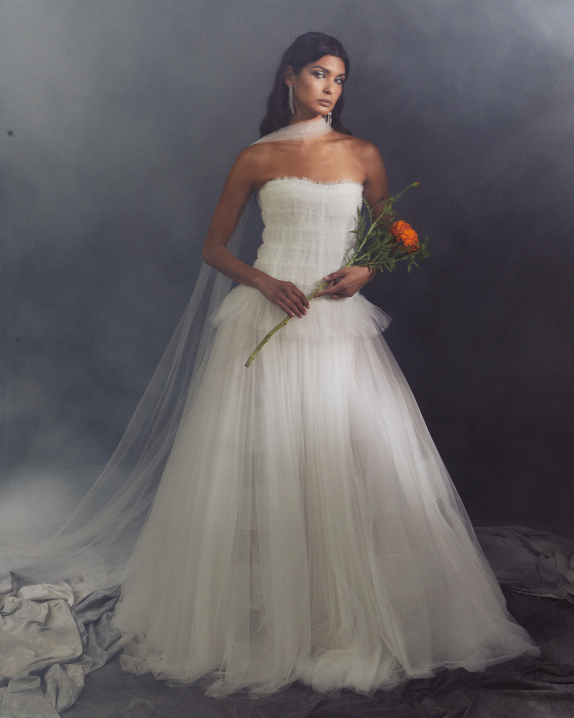 A woman in a strapless white wedding dress holds an orange flower while standing against a smoky, dark background.