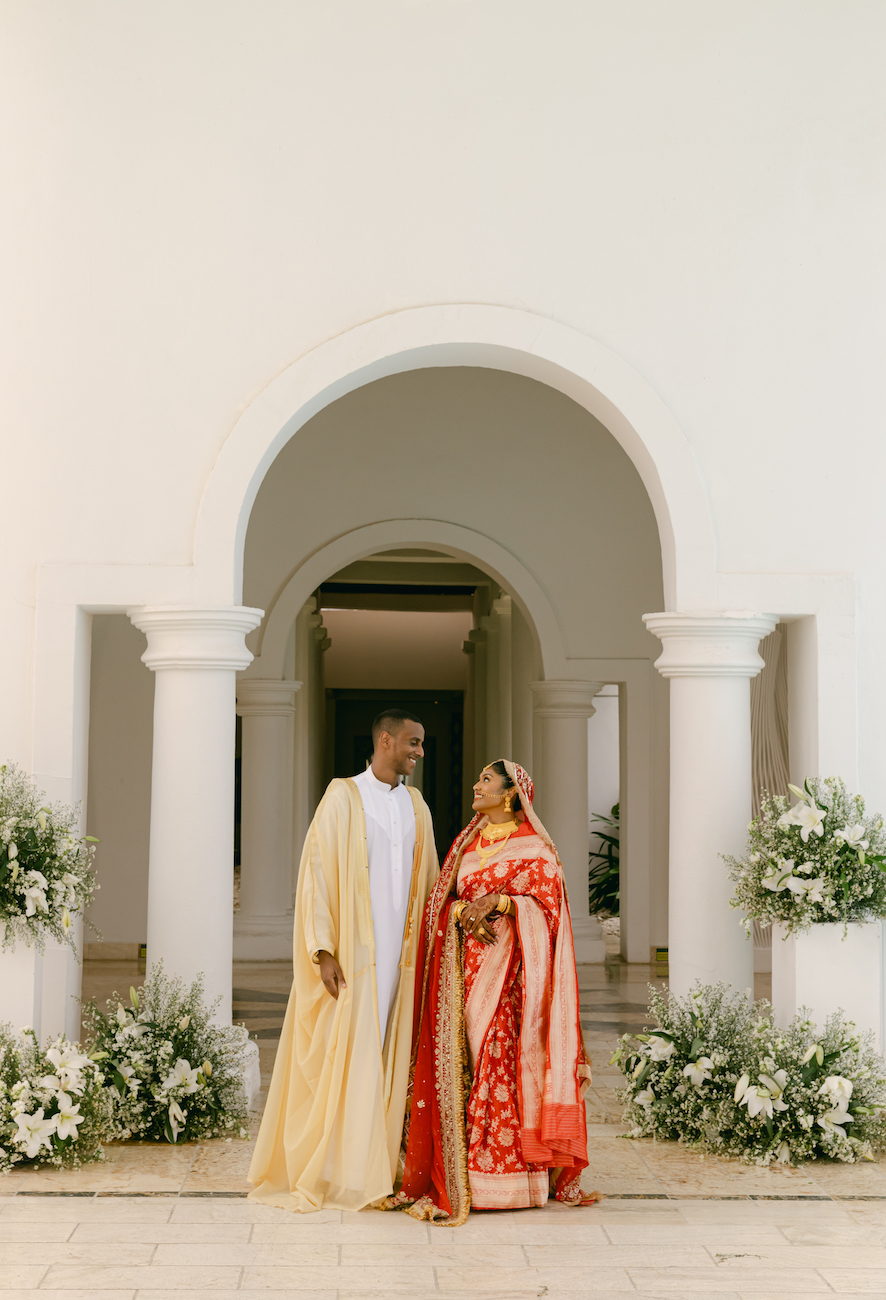 A couple in traditional attire stands in front of a white building with arched columns, surrounded by white floral arrangements.