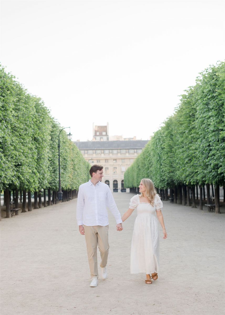 A couple walks hand in hand down a tree-lined path with a historic building in the background.