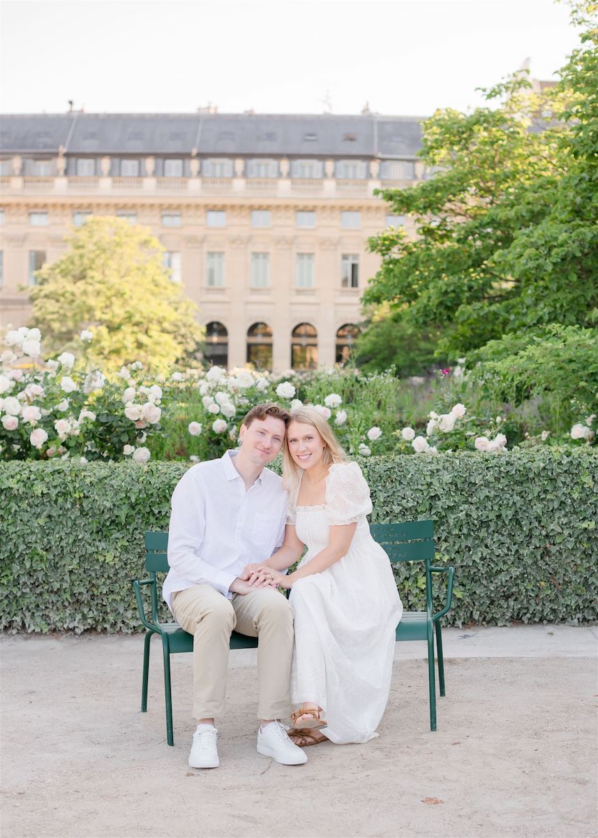 A couple sits together on a green bench in a garden, with blooming flowers and a historic building in the background.