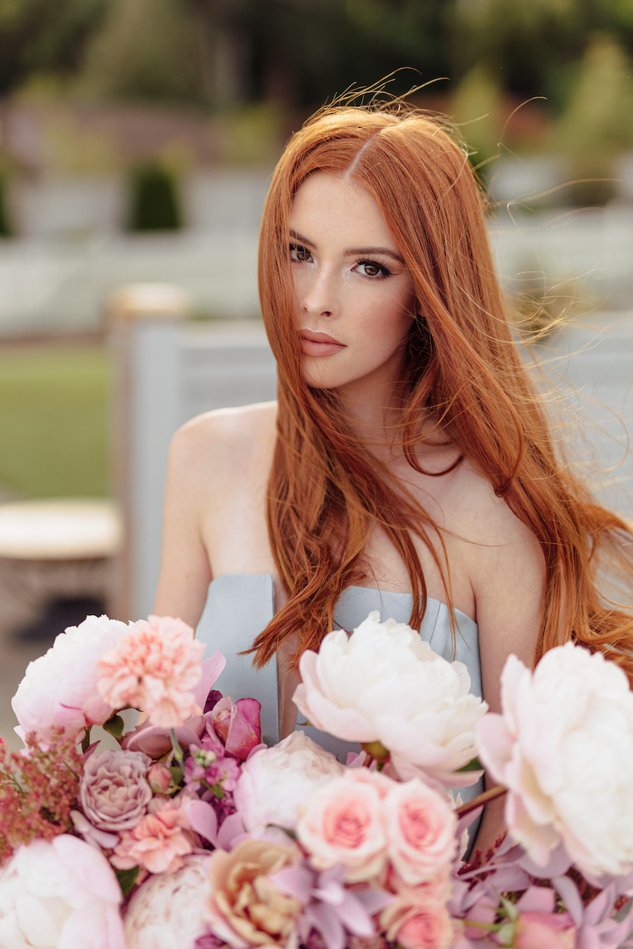 A woman with long red hair in a strapless dress holds a large bouquet of pink and white flowers outdoors.