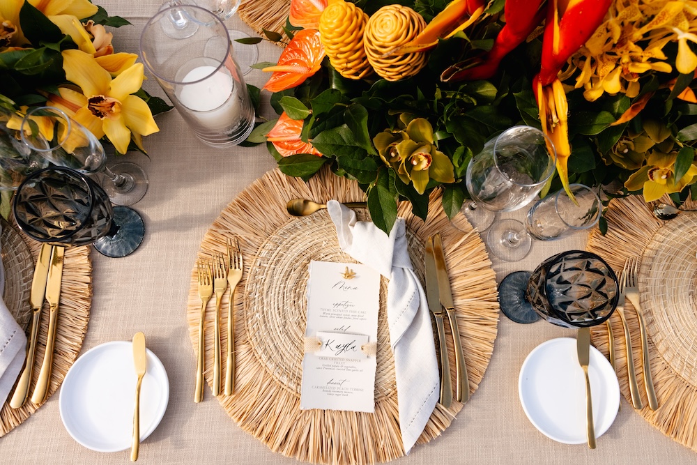 Table setting with woven placemats, white plates, gold cutlery, menus, napkins, wine glasses, and a centerpiece of yellow and orange flowers.