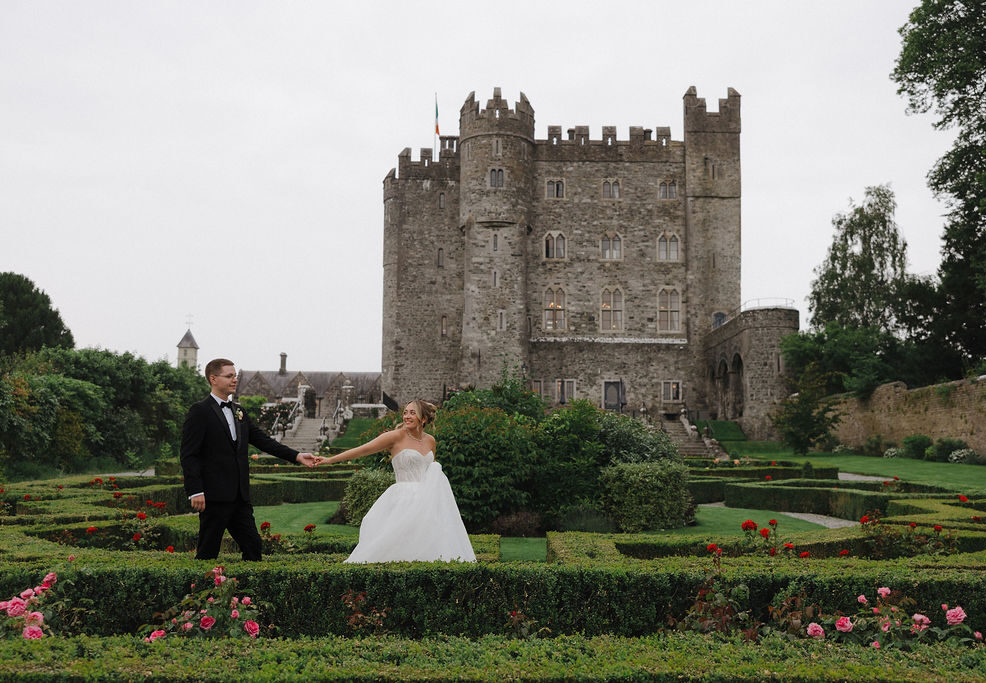 A bride and groom hold hands in a manicured garden with a large stone castle in the background.