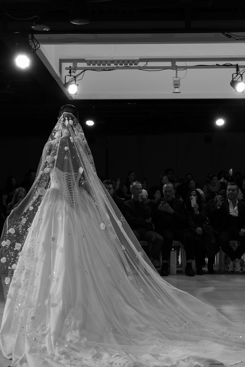 A bride in a long, embroidered veil and gown walks on a runway or stage, with an audience seated in the background.