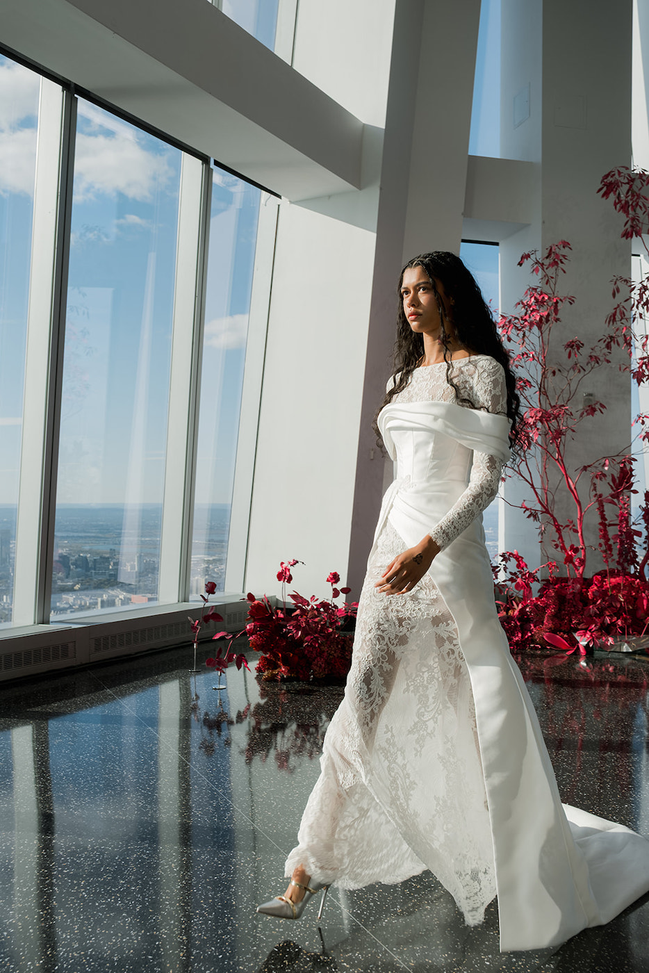 A woman in a long-sleeved white lace gown walks indoors by large windows, with red plants and cityscape visible in the background.