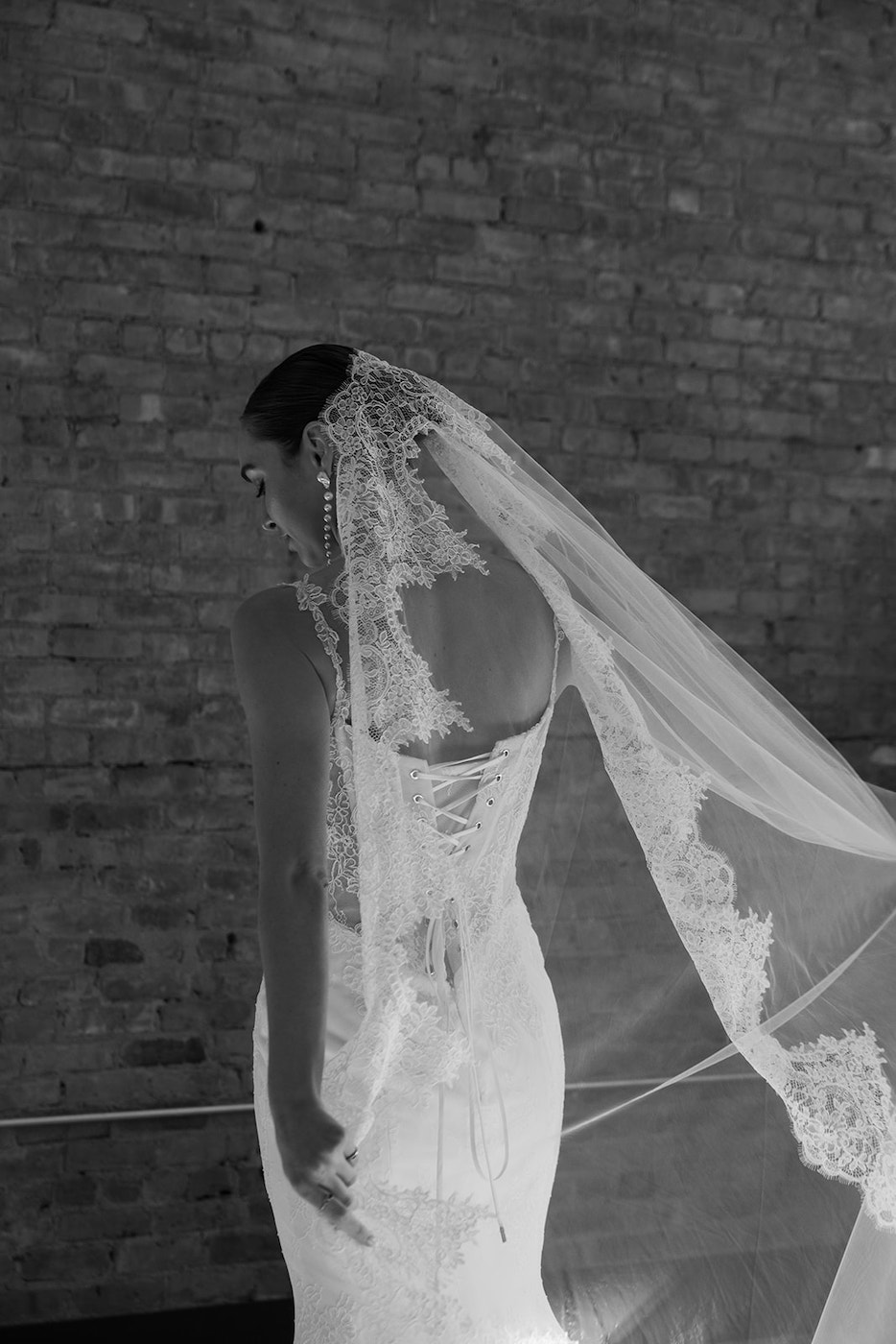 A bride in a lace wedding dress and veil stands against a brick wall, facing away from the camera.