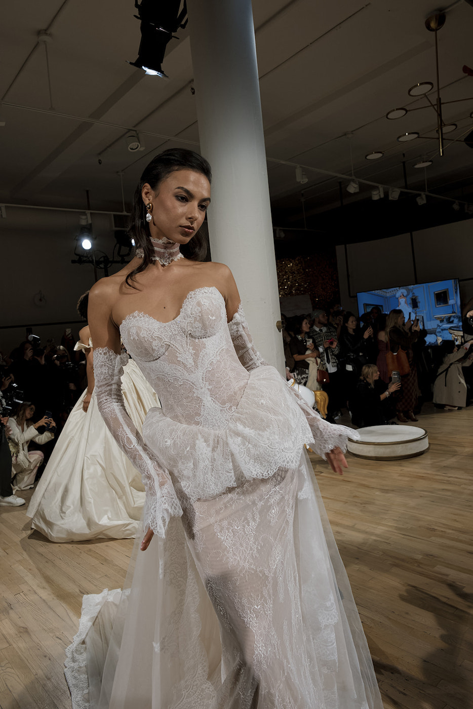 A model walks a runway wearing a strapless, fitted white lace gown with sheer panels, long gloves, and a peplum detail.