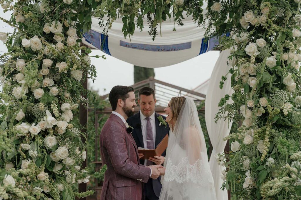A bride and groom stand under a floral arch, holding hands and smiling, while an officiant reads from a book during an outdoor wedding ceremony.