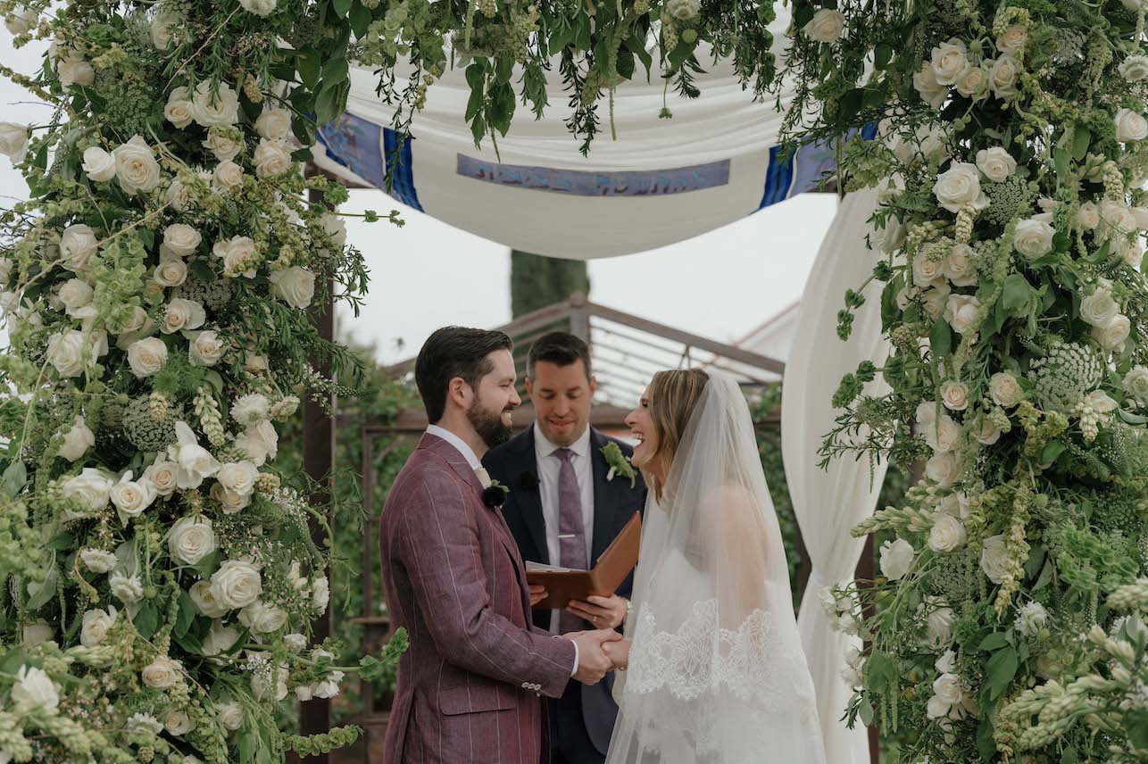 A bride and groom stand under a floral arch, holding hands and smiling, while an officiant reads from a book during an outdoor wedding ceremony.