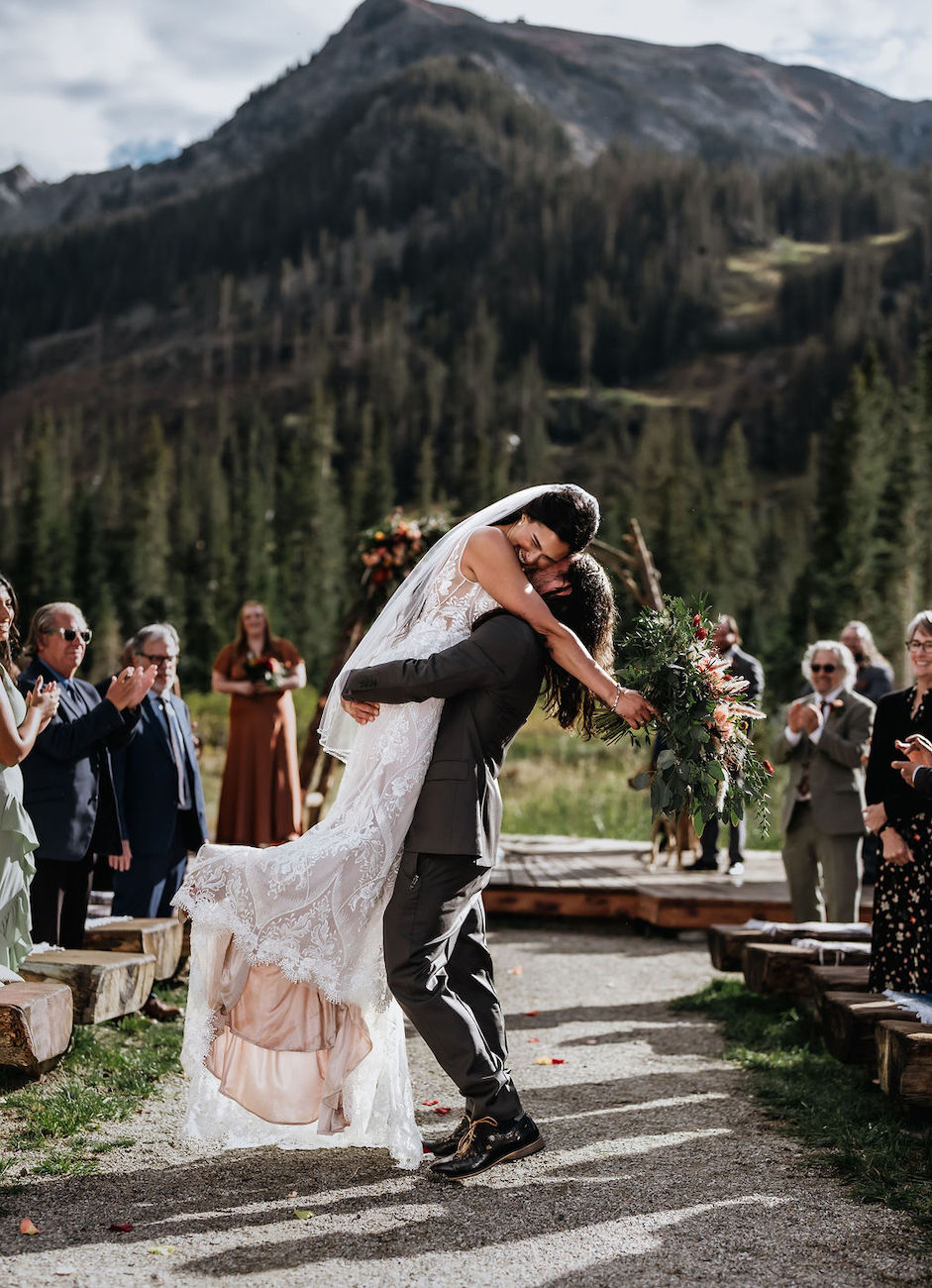 A groom lifts and embraces his bride outdoors on a stone path, surrounded by guests and trees with mountains in the background.